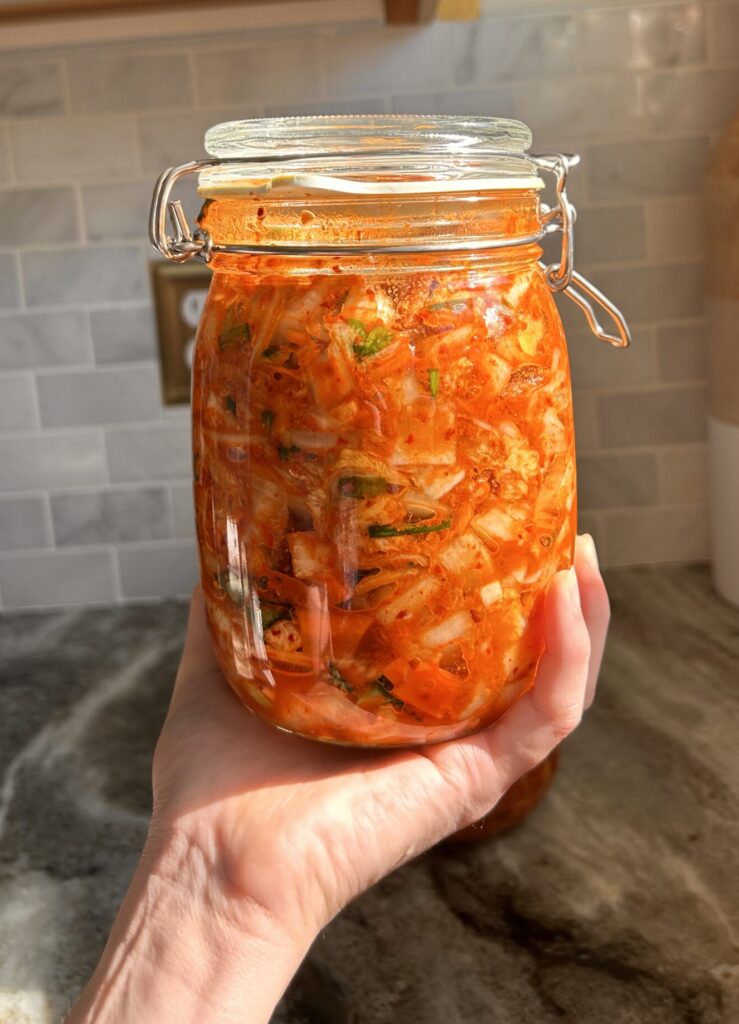 A jar of freshly fermented kimchi held up against a soft, neutral background, emphasizing its homemade quality.