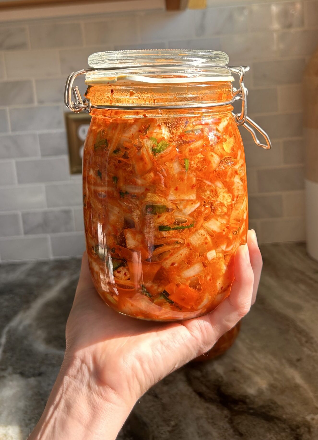 A jar of freshly fermented kimchi held up against a soft, neutral background, emphasizing its homemade quality.