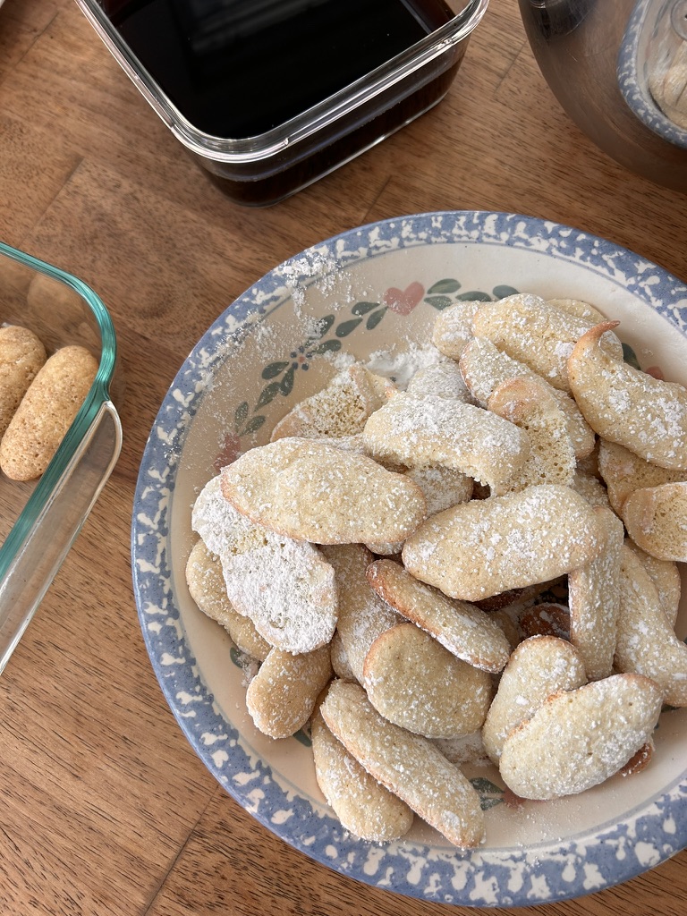 A bowl filled with homemade ladyfingers, dusted with powdered sugar, ready for the next step in a tiramisu recipe.