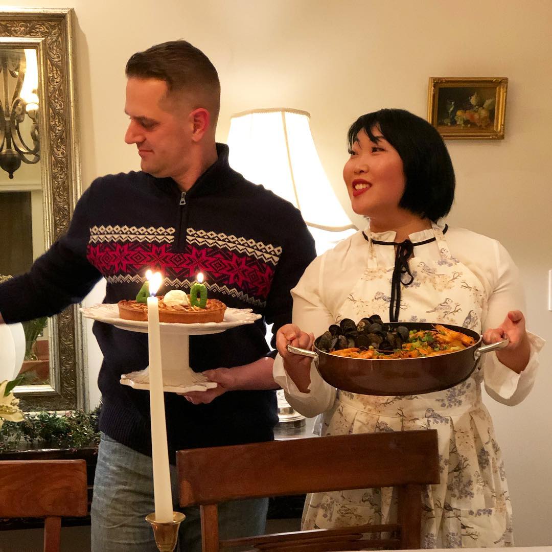 A woman in a patterned apron smiles while holding a large pot of seafood paella, standing next to a man holding a dessert plate with candles shaped as the number 40. They are in a warmly lit dining room, ready to serve the meal.