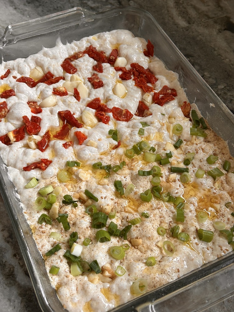 Sundried tomatoes and garlic cloves nestled into bubbly focaccia dough, drizzled with olive oil. Unbaked focaccia dough in a glass dish, topped with vibrant sundried tomatoes, whole garlic cloves, scallions, and sesame seeds