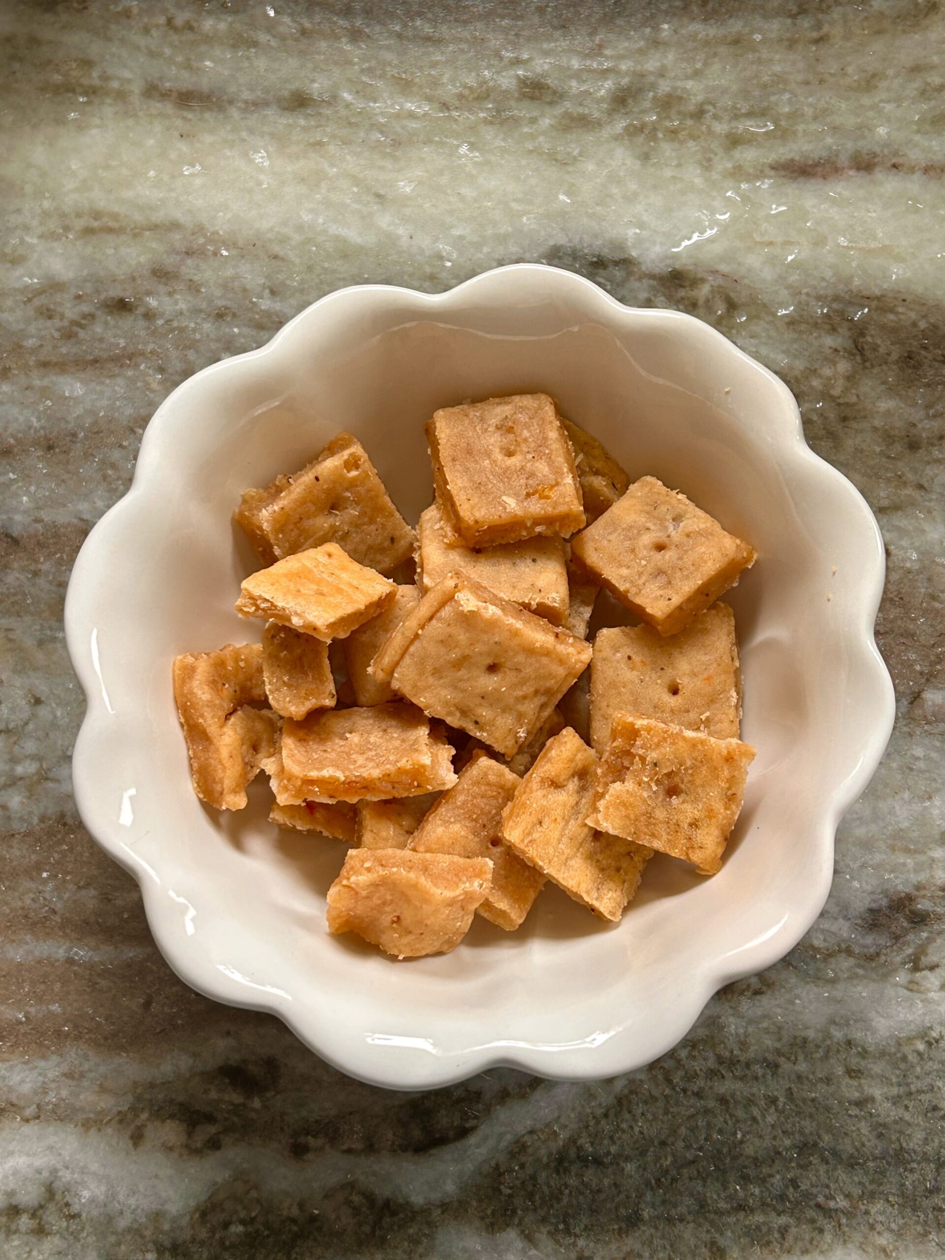 A small bowl of homemade sourdough cheddar crackers, golden and crispy, sitting on a marble counter.