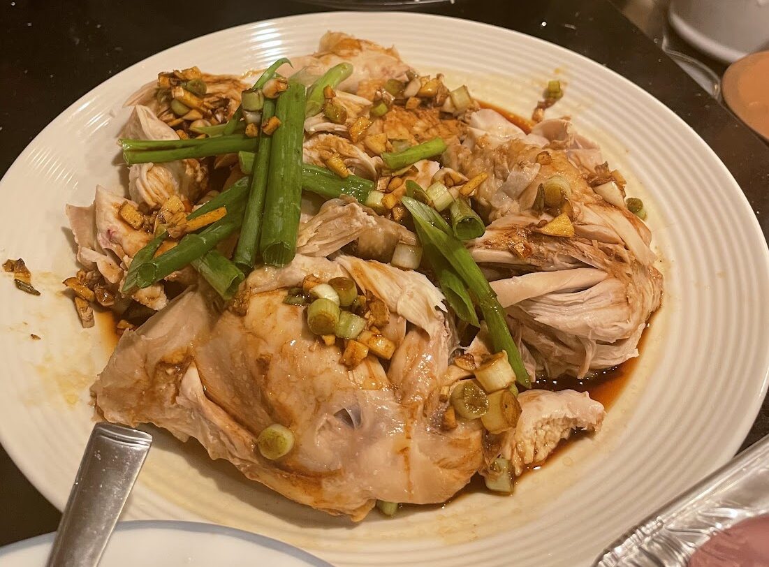 A plate of Hainanese chicken garnished with sliced green onions and crispy garlic, with fresh greens and enoki mushrooms in the background on a kitchen counter.