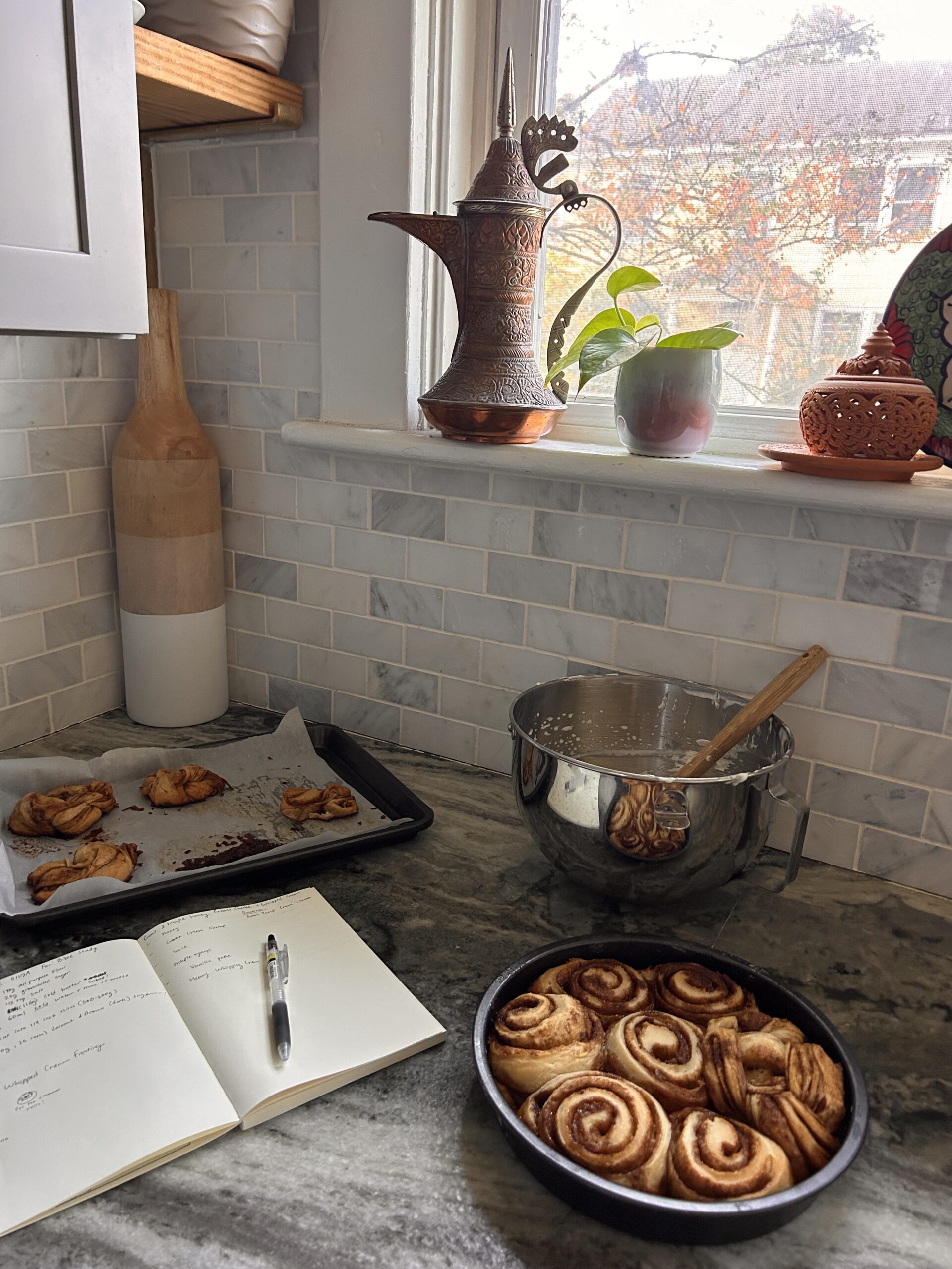 A pan of frosted cinnamon rolls held in a cozy kitchen setting with fall decor in the background. Title: Cinnamon Rolls in a Festive Kitchen Caption: Warm cinnamon rolls with brown butter frosting, surrounded by cozy fall decor. Description: A cozy kitchen setting featuring frosted cinnamon rolls, the perfect backdrop for fall baking. Pumpkin-themed decor and rustic touches create a festive atmosphere.