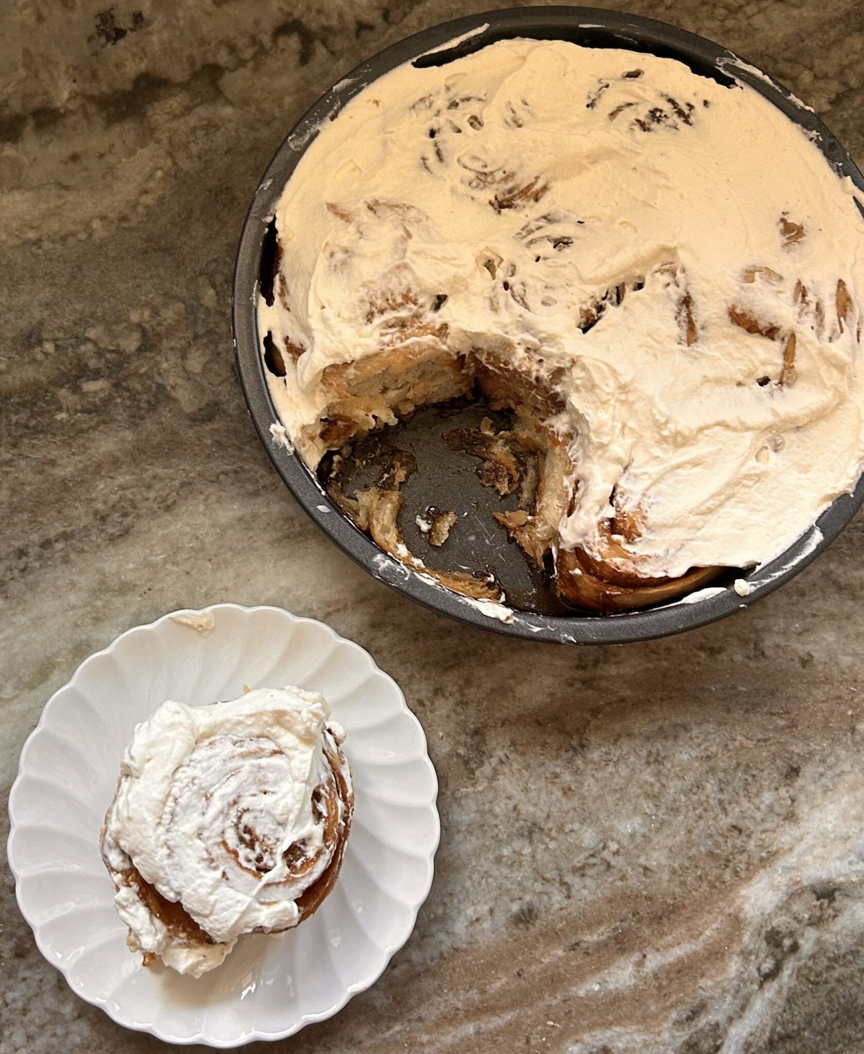 A frosted cinnamon roll on a white plate next to a pan of frosted rolls with a slice missing.