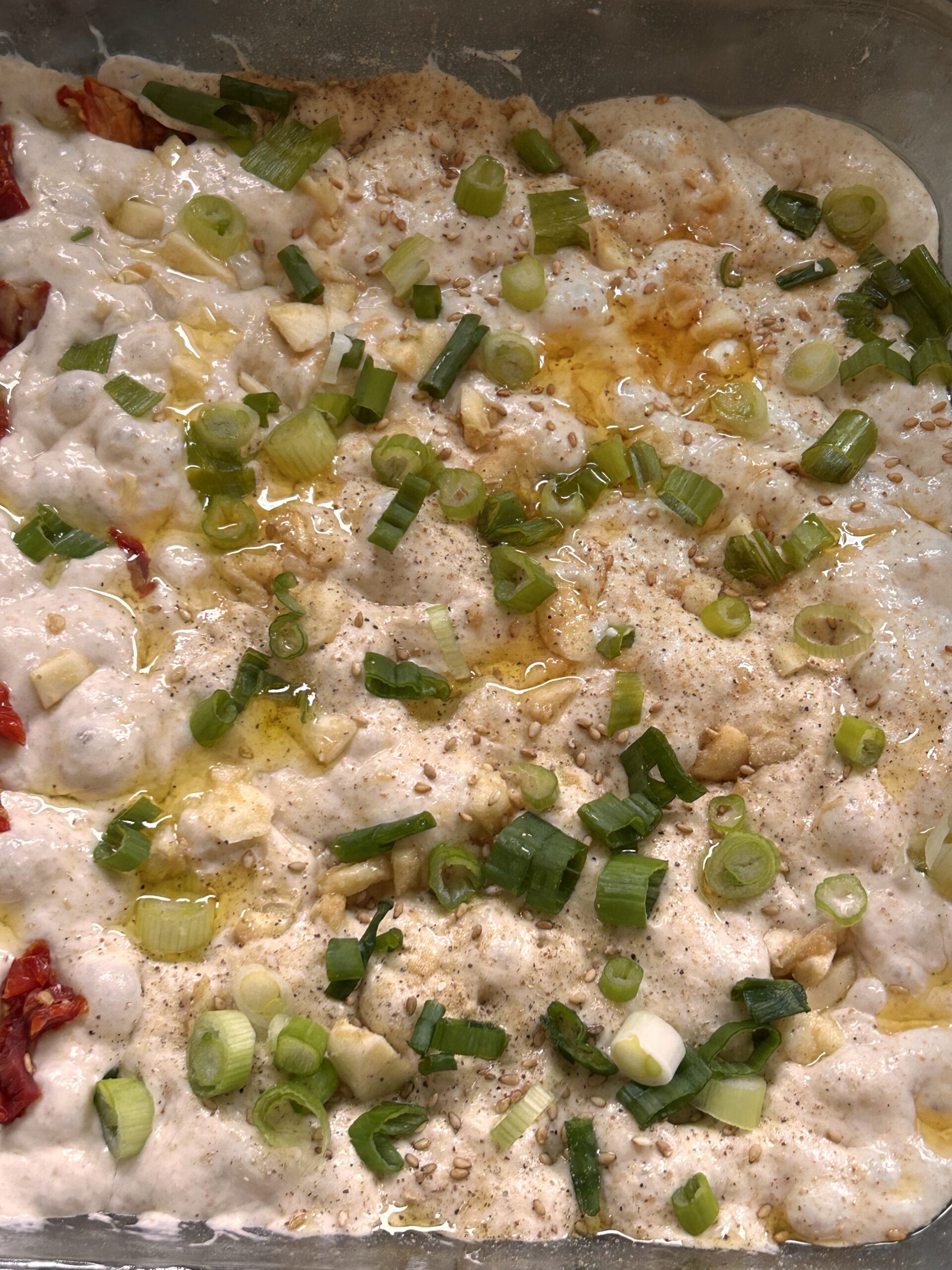 A bubbly focaccia dough in a glass dish, ready to be topped with flavorful ingredients.Close-up of freshly seasoned focaccia topped with scallions, sesame seeds, garlic, and miso paste.