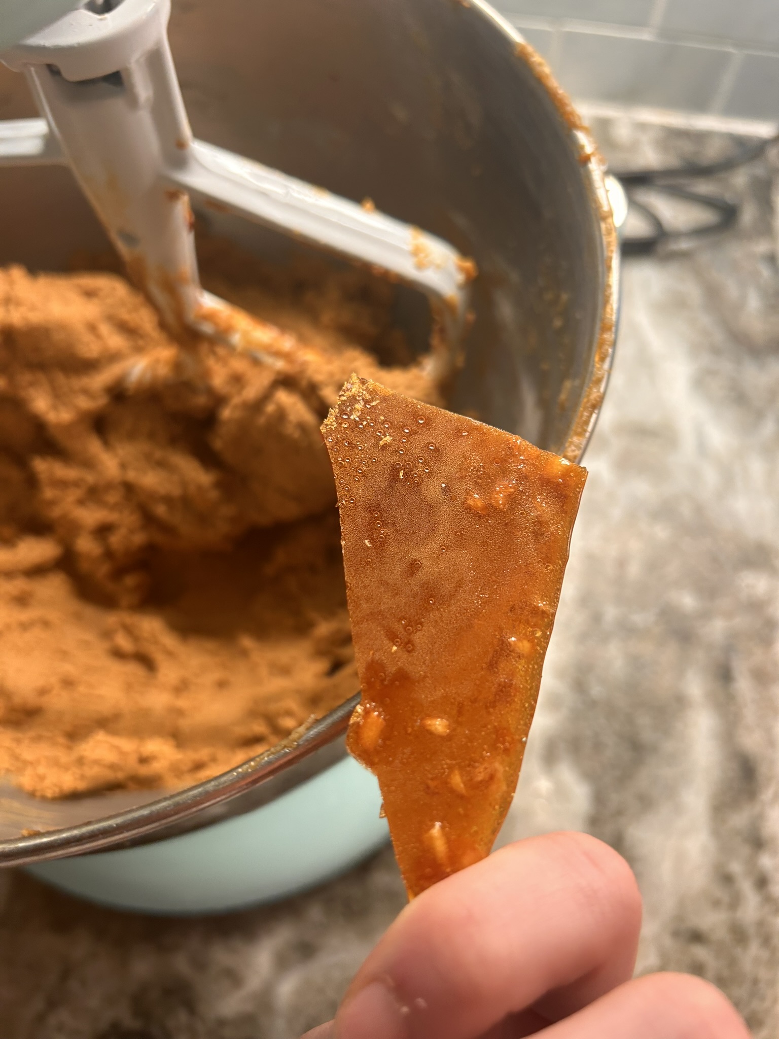 A caramelized burnt ginger sugar shard held over a bowl of ginger cookie dough, showcasing the vibrant amber color of the sugar.