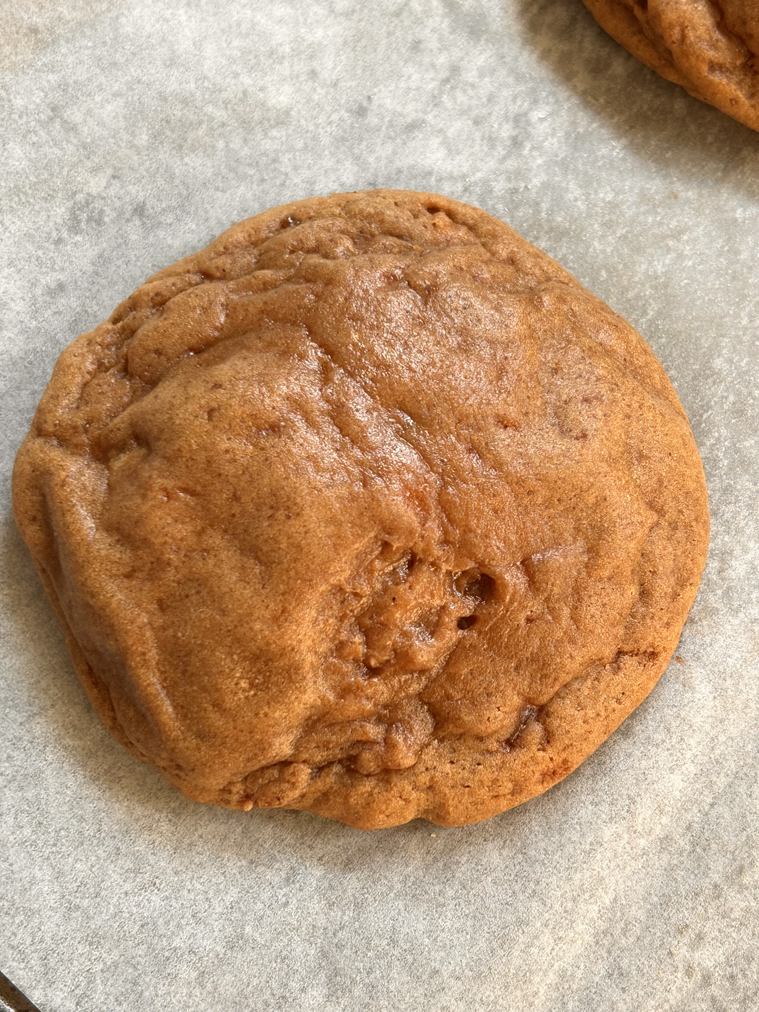 A close-up of a freshly baked burnt sugar ginger cookie with a golden-brown, slightly cracked surface, sitting on parchment paper.