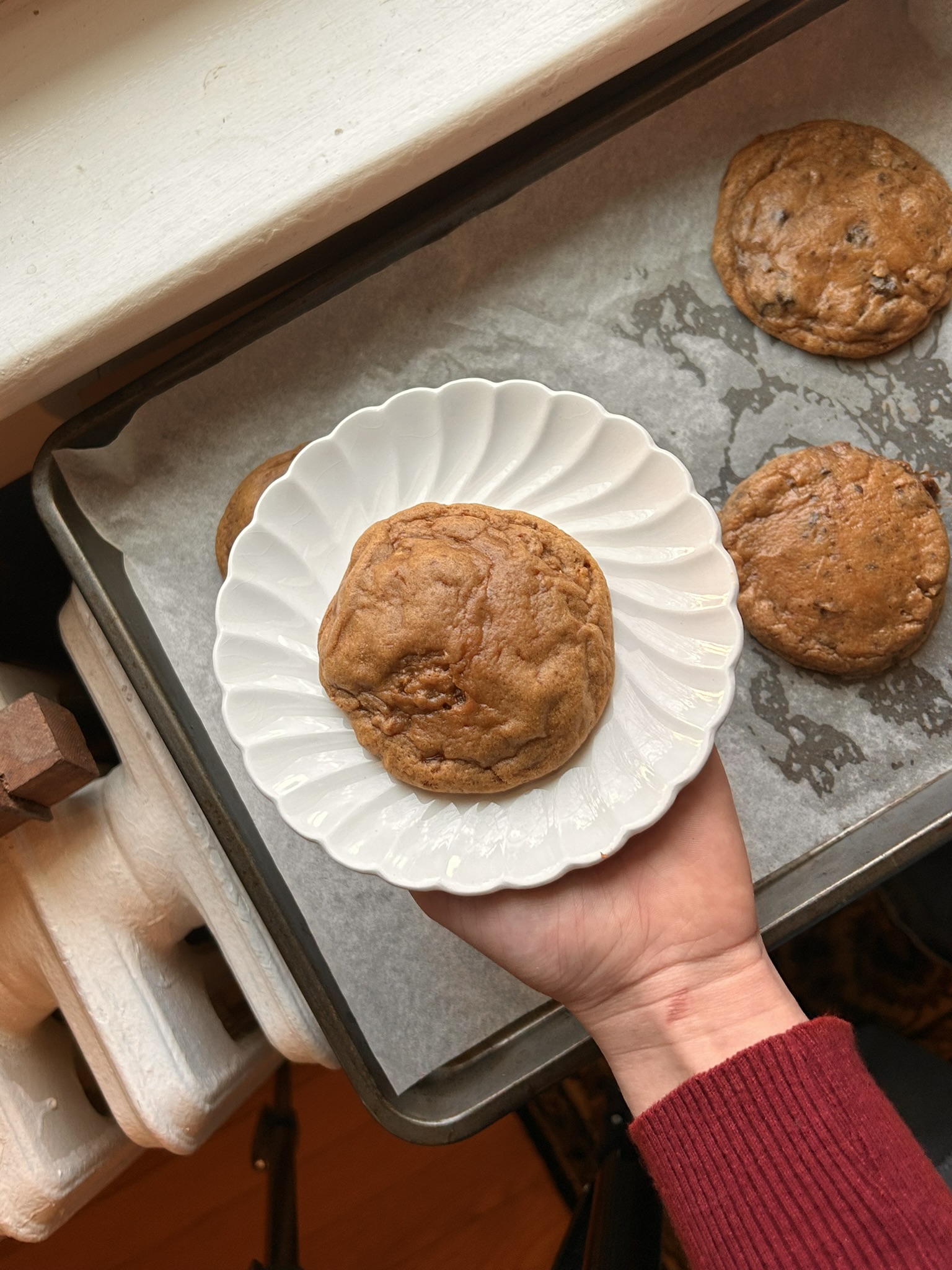 A woman in a red sweater holding a white plate with a golden-brown burnt sugar ginger cookie, smiling warmly in a cozy kitchenA tray of freshly baked burnt sugar ginger cookies on parchment paper, with warm golden-brown tops glistening in natural light