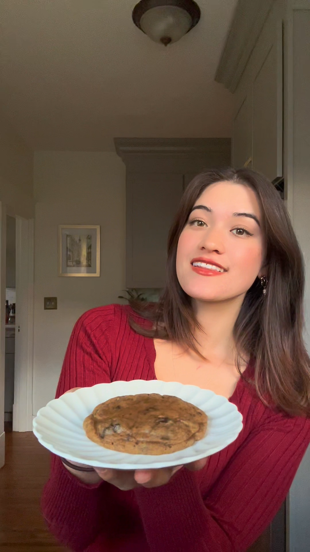 A smiling woman holding a white plate with a freshly baked brown butter miso chocolate chunk cookie, showcasing its golden-brown texture and chocolate chunks.