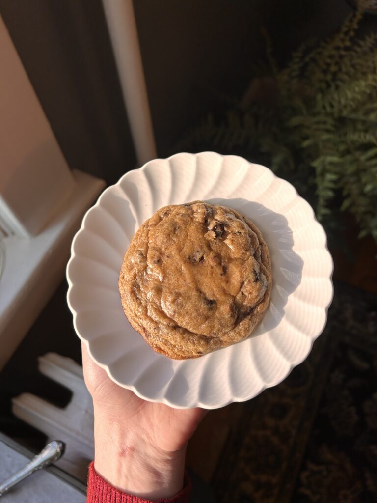 A single brown butter miso chocolate chunk cookie on a scalloped white plate, placed near a sunny window.