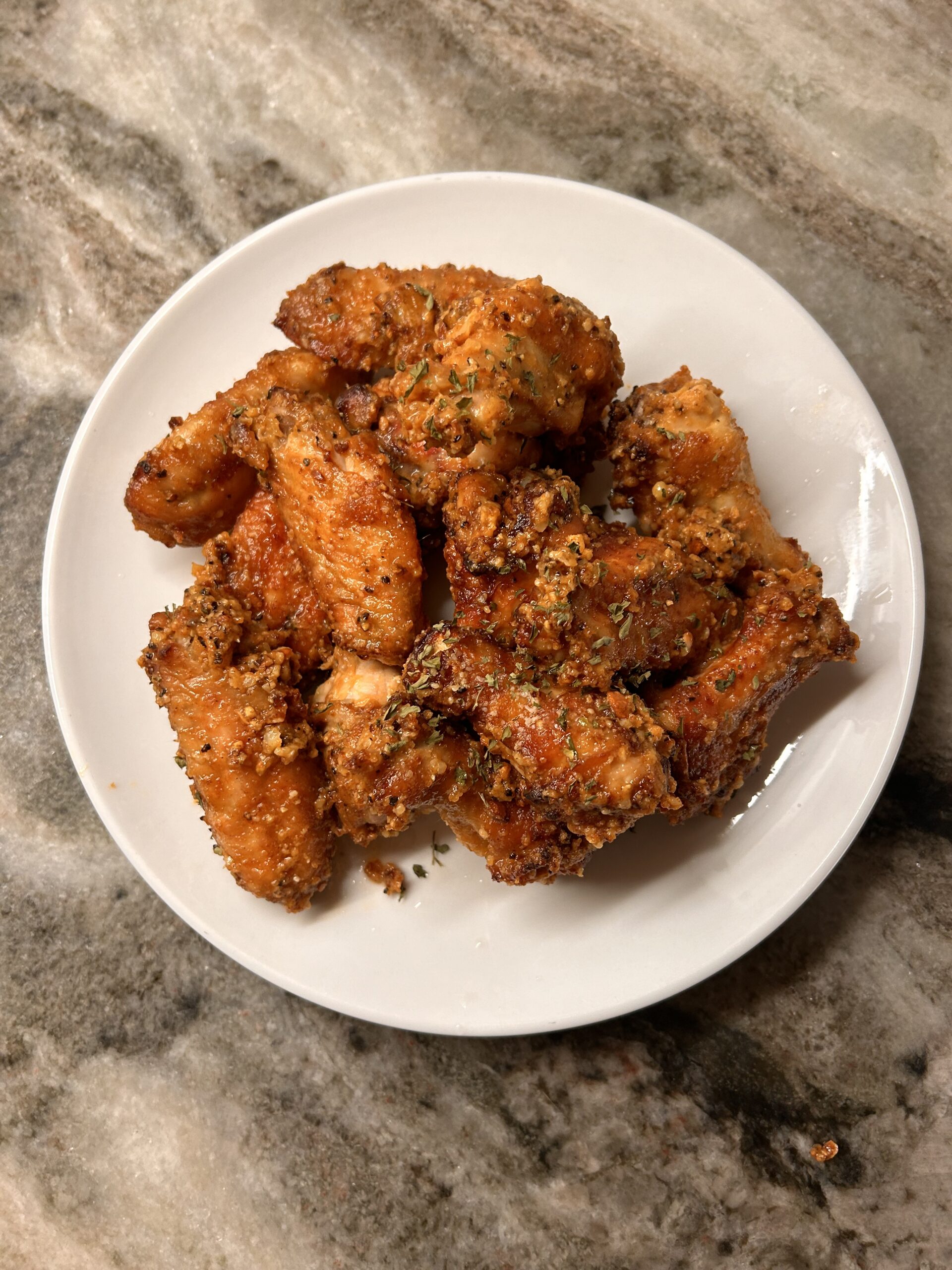 Baked Lemon Pepper Chicken Wings These baked lemon pepper wings bake up golden and crispy on the outside while staying juicy inside. Top-down view of crispy baked chicken wings with lemon pepper seasoning, garlic, and herbs on a round white plate against a marbled kitchen counter background