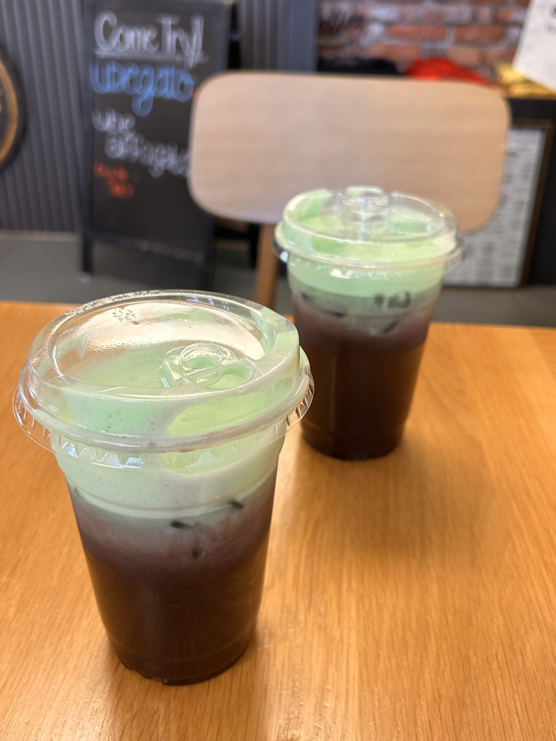 Two lattes topped with vibrant green foam sitting on a wooden café table with a chalkboard sign in the background.