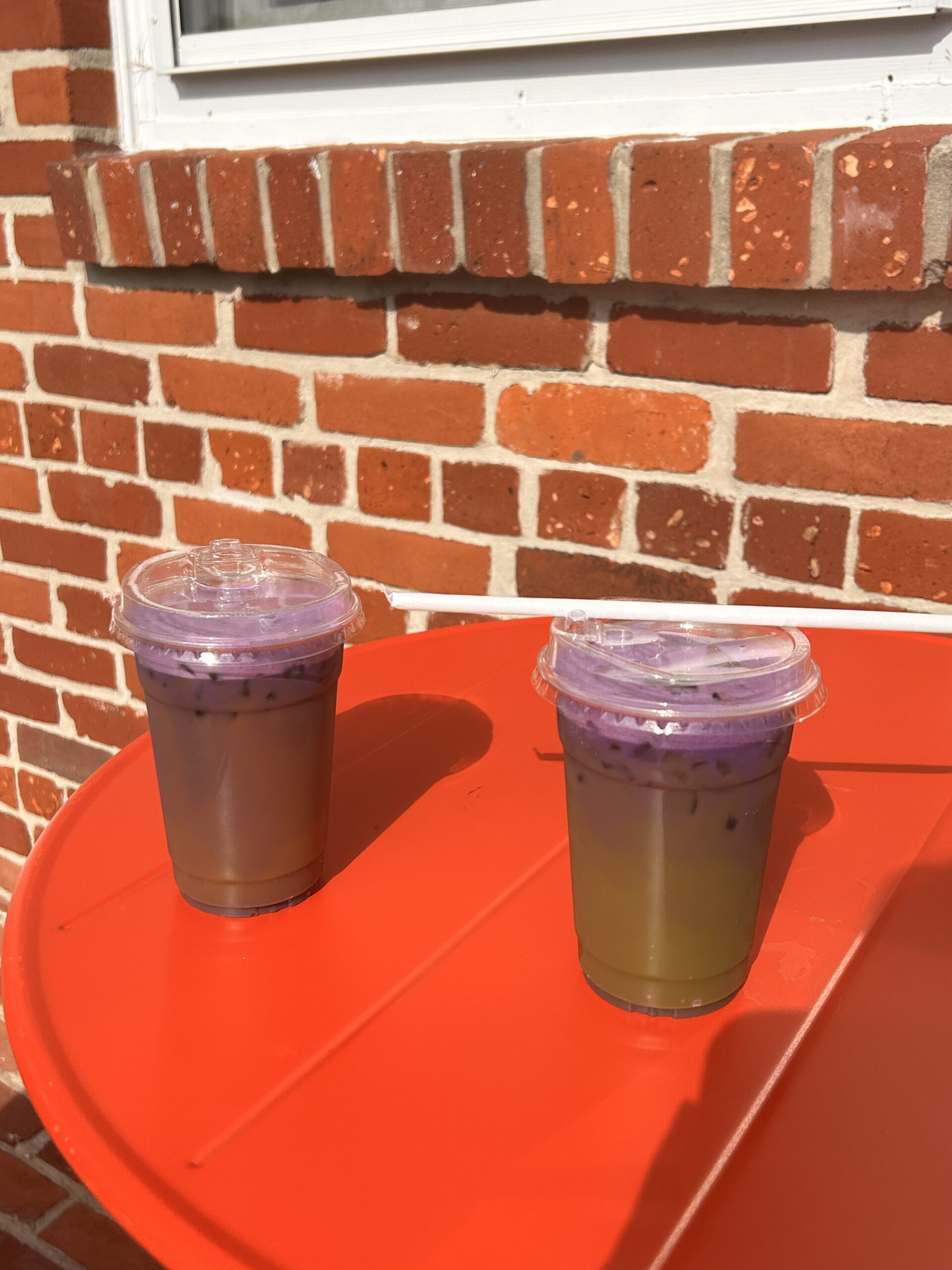 Two drinks on a red table outside with bright natural light and a brick wall backdrop, each topped with ube cold foam.