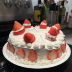 A Christmas strawberry shortcake with layers of light yellow sponge cake, fluffy whipped cream, and glossy red strawberries. Garnished with additional strawberry slices and whipped cream on top, the dessert’s red and white colors look especially festive for the holiday table.