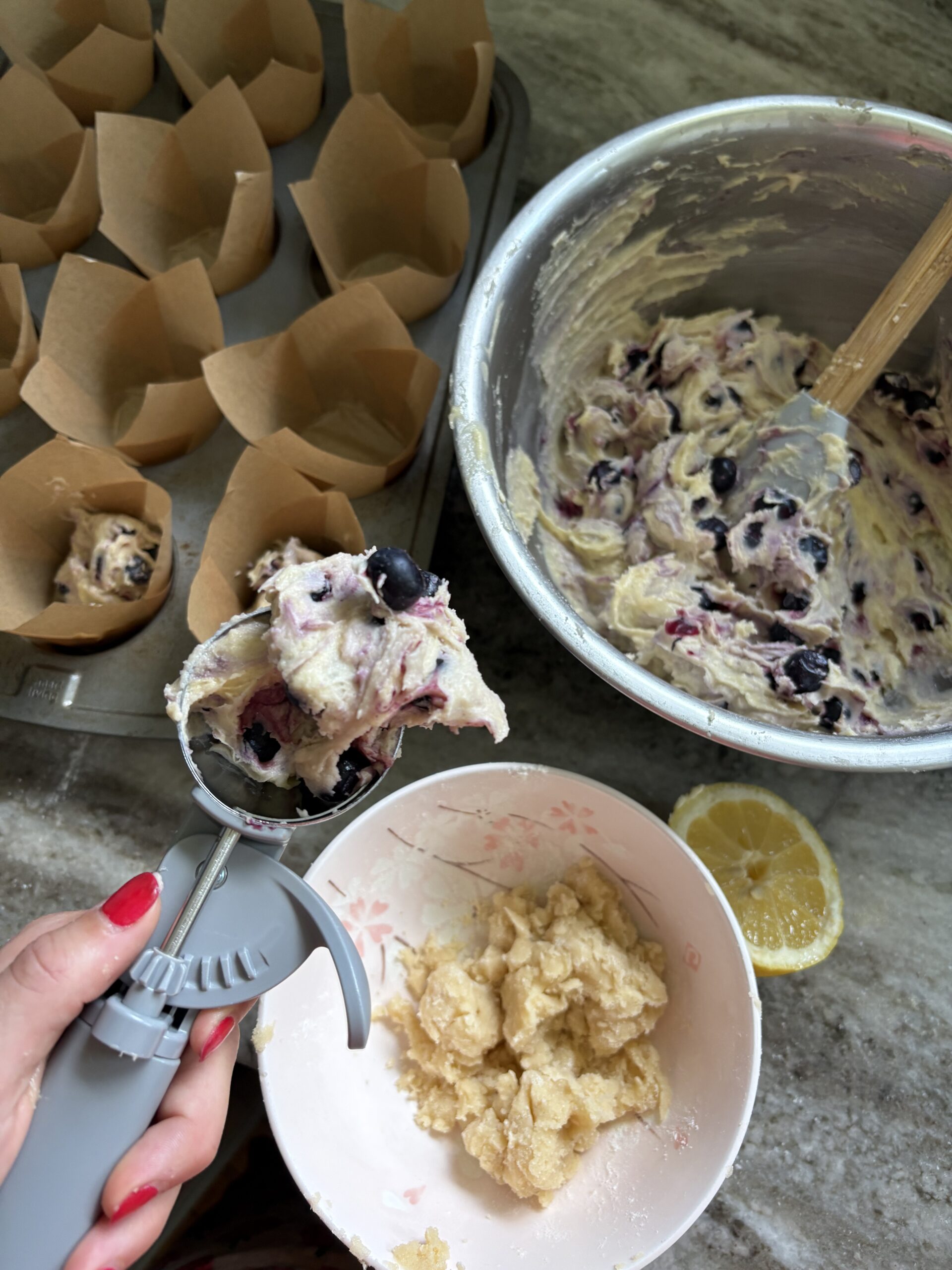 Hand scooping blueberry muffin batter into tulip muffin liners next to cinnamon streusel topping